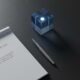 A top-down view of a clean desk with a simple planner, a fountain pen, and a bright blue glass paperweight.Featured Image Description: The scene is an overhead surveillance view of a minimalist workspace. A heavy, dark slate desk holds a clean white planner open to a fresh page. A silver fountain pen lies perfectly straight beside it. A single, solid glass cube glows with a soft electric blue light from within, symbolizing the clarity and focus of the top performers. The environment is silent and orderly, representing the calm before the busy week.Featured Image Title: The Order of Elite Business Starts