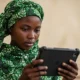 A young Nigerian woman in a green headscarf focused on a digital tablet, illustrating the hardware requirements for Secure Electronic Voting in Nigeria.