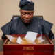 A close-up of a Nigerian official looking at Flood Water in the Governor’s Inbox as official documents float in murky water on his desk.