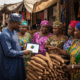 A government official using a digital tablet for community sentiment analysis for government while listening to traders in a busy Nigerian market.