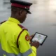 A Nigerian traffic warden in a yellow and maroon uniform checking a digital tablet for road data while Using Cameras to Watch Markets on a wet tarmac.