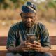 Nigerian citizen using digital engagement platform on smartphone showing state government feedback formFeatured Image Description:
Digital photograph of a middle-aged Nigerian man in casual clothing seated on a wooden bench outdoors, holding a smartphone displaying a state government citizen feedback interface. His expression shows concentration as he reads the screen. Natural daylight. Blurred background of a residential compound visible. The phone screen shows green and white government branding elements but no readable text. Taken in first quarter 2026.Featured Image Title:
digital-citizen-engagement-platform-nigeria-2026.jpg
