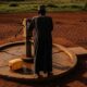 A person with hands on a broken borehole pump in a dry, rural Nigerian landscape.