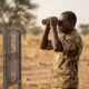 Nigerian soldiers at border post near Niger Republic looking across fence line after coup closureFeatured Image Description:
Digital photograph of Nigerian military personnel in camouflage uniforms standing at a border observation post along the Nigeria-Niger frontier. Soldiers use binoculars to scan the horizon. Dry savannah landscape stretches behind them. A faded border marker visible in foreground. Date stamp indicates dry season 2026.Featured Image Title:
niger-coup-border-security-nigeria-2026.jpg