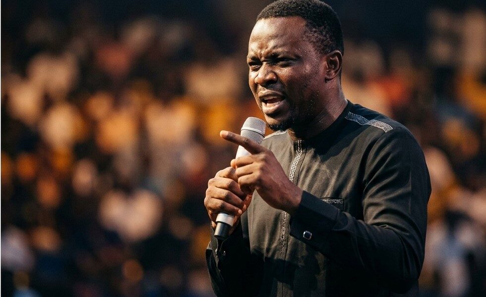 Nigerian comedian performing on stage at indoor arena with crowd visible in background and stage lights creating dramatic effectFeatured Image Description:
Digital photograph of a Nigerian comedian performing at a major comedy show in Lagos. The comedian stands center stage holding a wireless microphone, mouth open mid-performance. Stage lighting creates dramatic shadows. Audience visible in foreground as dark silhouettes with occasional phone screens glowing. Arena setting suggests capacity over 3,000 attendees. Professional production values visible in lighting rig and sound equipment. Date indicates 2026 performance.Featured Image Title:
nigerian-comedian-live-performance-arena-2026.jpg