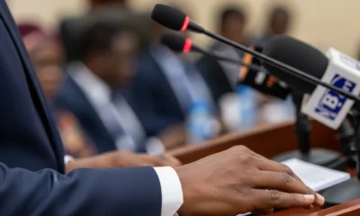 Hand adjusts mic at podium; Nigerian flag blurred.