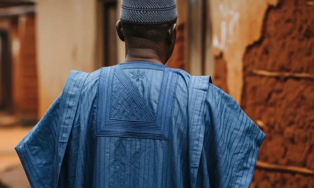 Man in agbada stands before old mud house