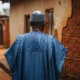 Man in agbada stands before old mud house
