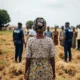 Elderly woman in field; NDLEA figures blur behind.