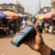 A hand holds a black mobile payment terminal against a blurred outdoor market background