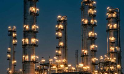 Silhouetted industrial towers of a refinery against a dark blue twilight sky with glowing lights