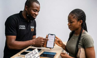 A Nigerian man and woman stand at a wooden counter where the man points to the battery health screen on a blue iPhone 13. A small sign on the desk lists the price for a refurbished 128GB model between N420,000 and N480,000.