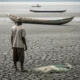 Fisherman stands on cracked mud with canoe in distance.
