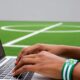 Hands type on laptop with Sporting Lagos wristband. Football pitch background.